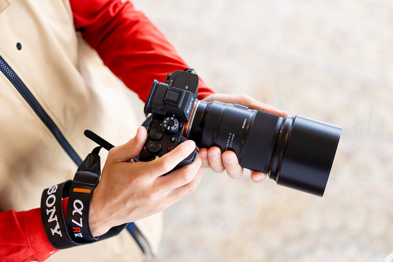 Photographer holding a DSLR camera with telephoto lens, wearing a red jacket and camera strap.