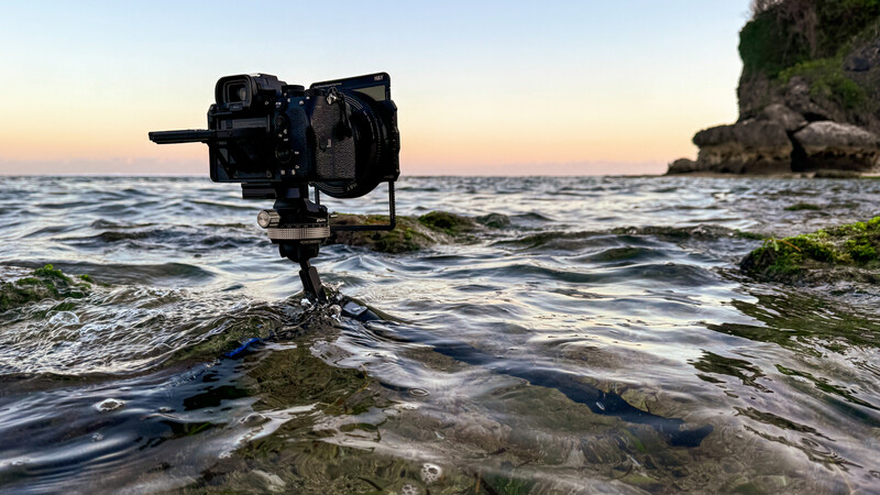 Professional video camera mounted on tripod in shallow coastal water at sunset, with rocky shore and waves in foreground.