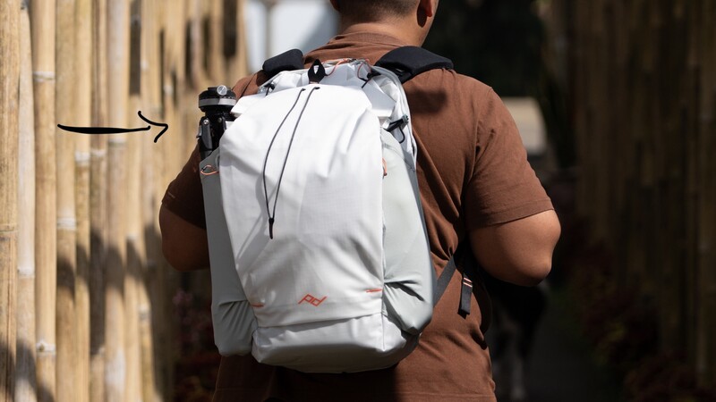 Person wearing white roll-top backpack over brown shirt, photographed from behind on a sunny street.
