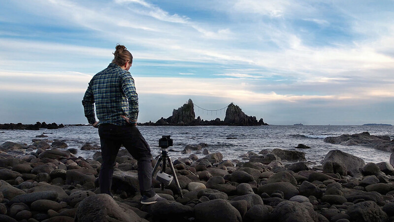 Photographer standing on rocky shore with camera on tripod, facing dramatic rock formations in ocean at dusk.