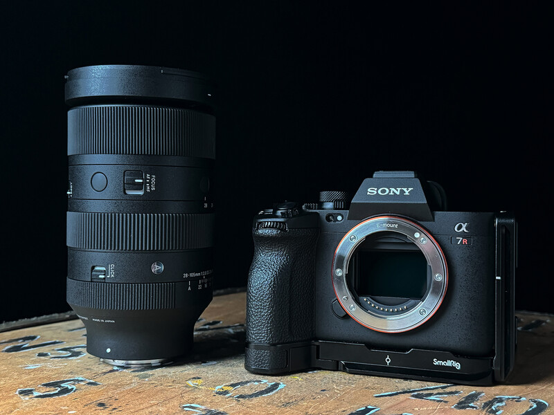 Sony a7 IV mirrorless camera body with telephoto lens and battery grip on dark background.