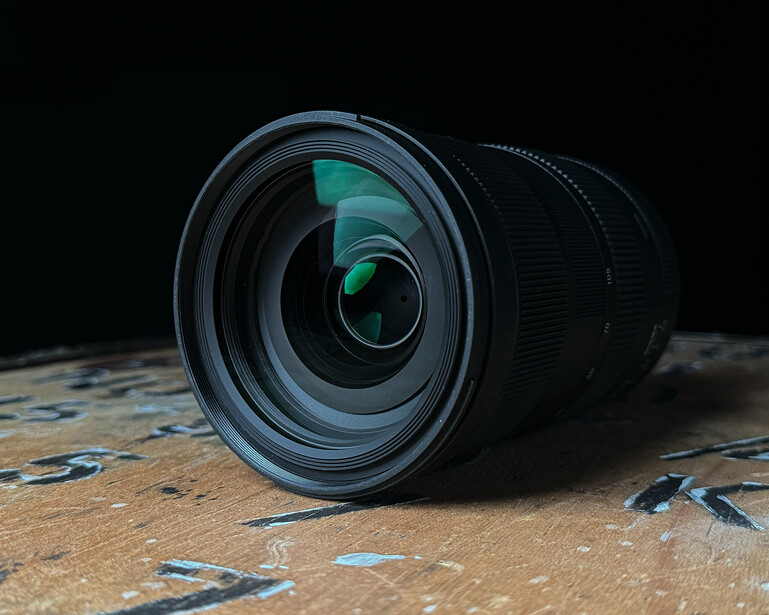 Wide-angle zoom lens positioned on a textured surface against dark background, showing front glass elements and aperture blades.