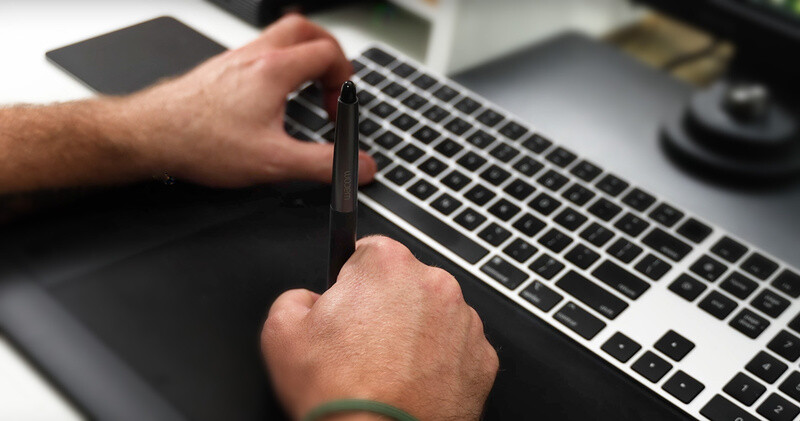 Hands holding a stylus over a laptop keyboard and trackpad during a creative work session.