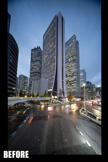 Urban street at dusk with illuminated skyscrapers and wet pavement reflecting city lights.