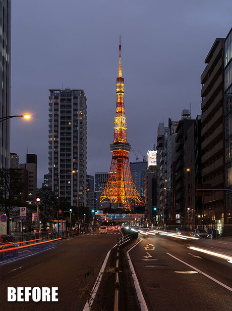 Illuminated red and gold tower at dusk framed by urban buildings with car light trails on the street.