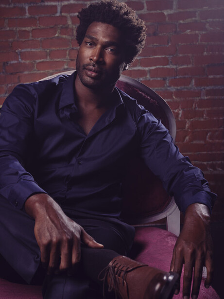 Man in dark blue shirt seated against red brick wall in studio lighting.
