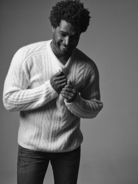 Man in white cable-knit sweater and dark jeans smiling while adjusting his collar against gray background.