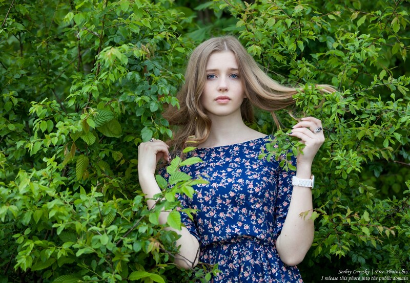 Young woman in navy floral dress posing against dense green foliage.