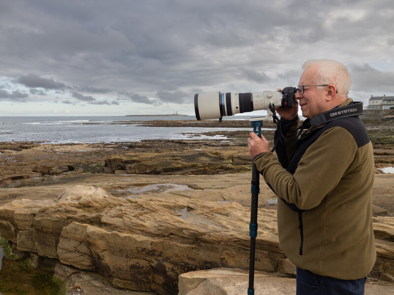 Older man with glasses photographing coastal landscape using telephoto lens on tripod.