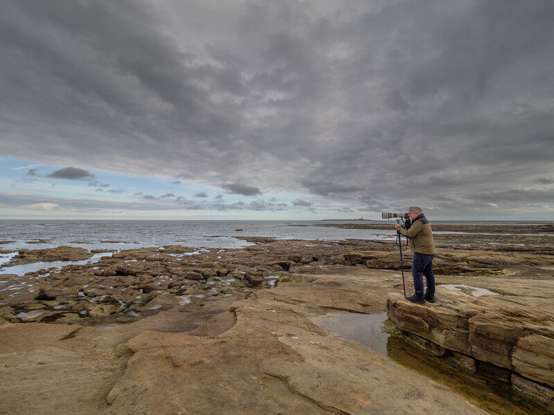 Photographer on rocky outcrop using tripod-mounted telephoto lens, facing expansive tidal beach under dramatic cloudy sky.
