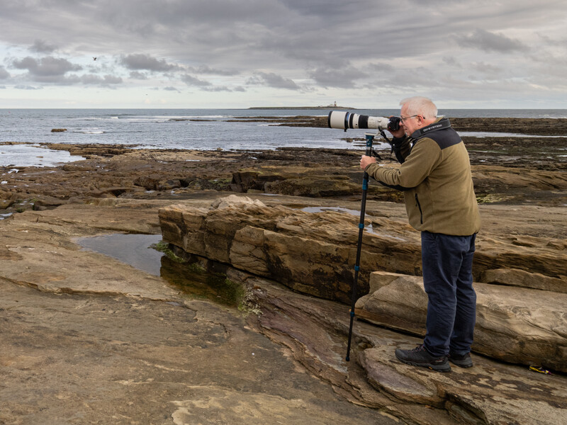 Photographer with telephoto lens on rocky coastal beach under overcast sky.