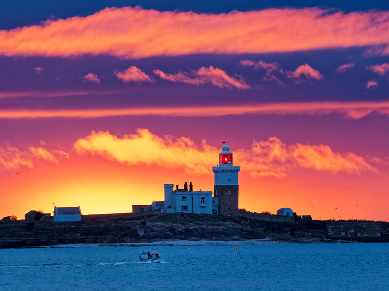 Silhouetted lighthouse on rocky coastline against vibrant orange and purple sunset sky with golden clouds.