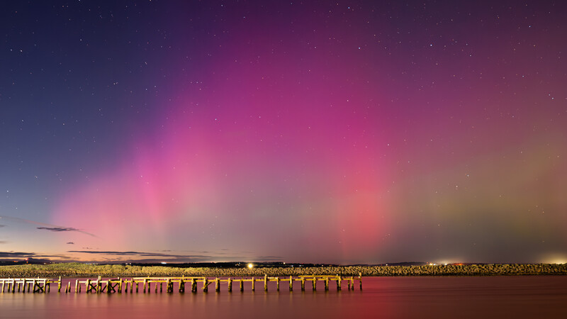 Vibrant aurora-like lights illuminate the sky above a waterfront pier at dusk.