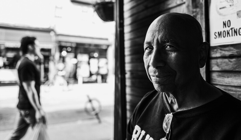 Bald man in dark shirt looking toward camera with blurred urban street scene behind him in black and white photography.