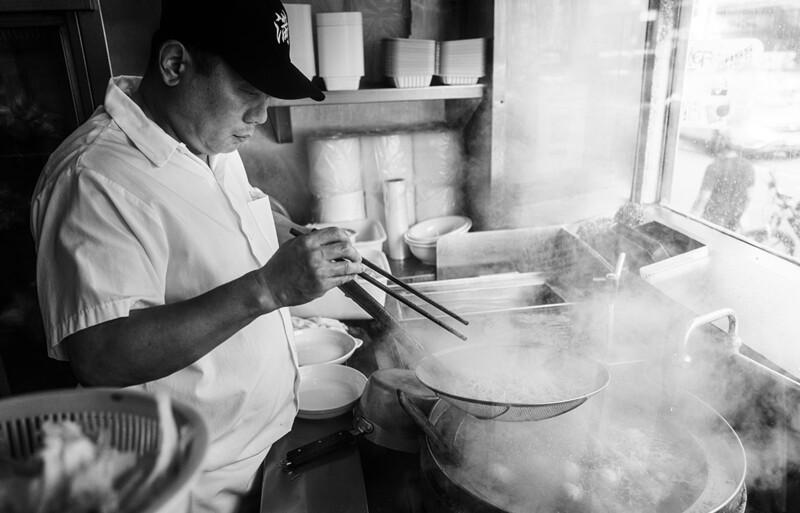 Cook stirring large pot of steaming liquid with chopsticks in a professional kitchen.
