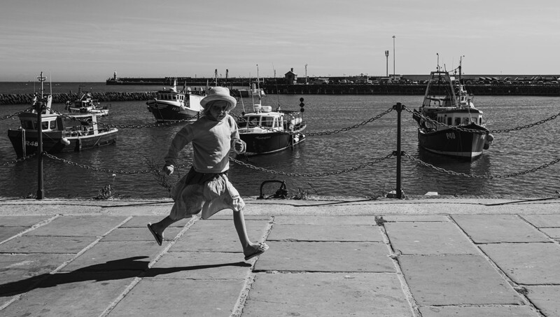 Person in motion running across a waterfront dock with fishing boats anchored in the harbor behind.