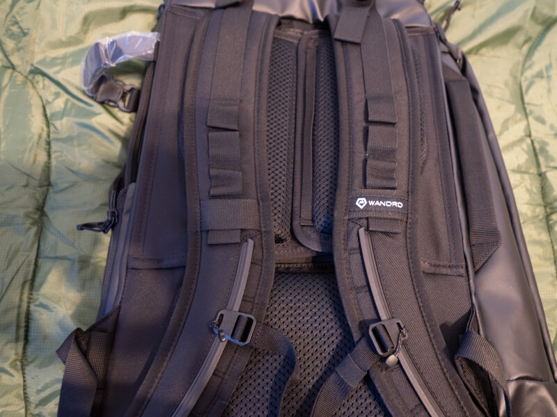 Back view of a dark gray camera backpack with padded straps and mesh ventilation panels displayed on a patterned fabric surface.