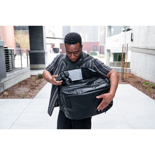 Photographer examining a black camera bag outdoors on a rooftop with city buildings visible in the background.