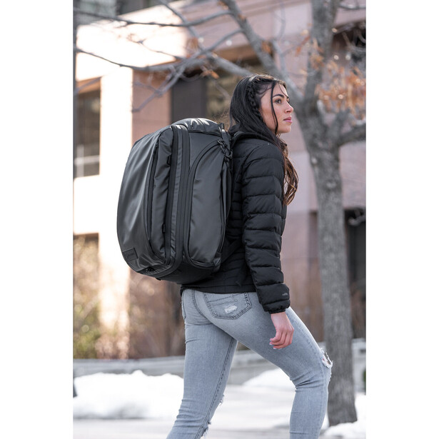 Person wearing black puffer jacket and light blue jeans carries large black backpack in urban setting.