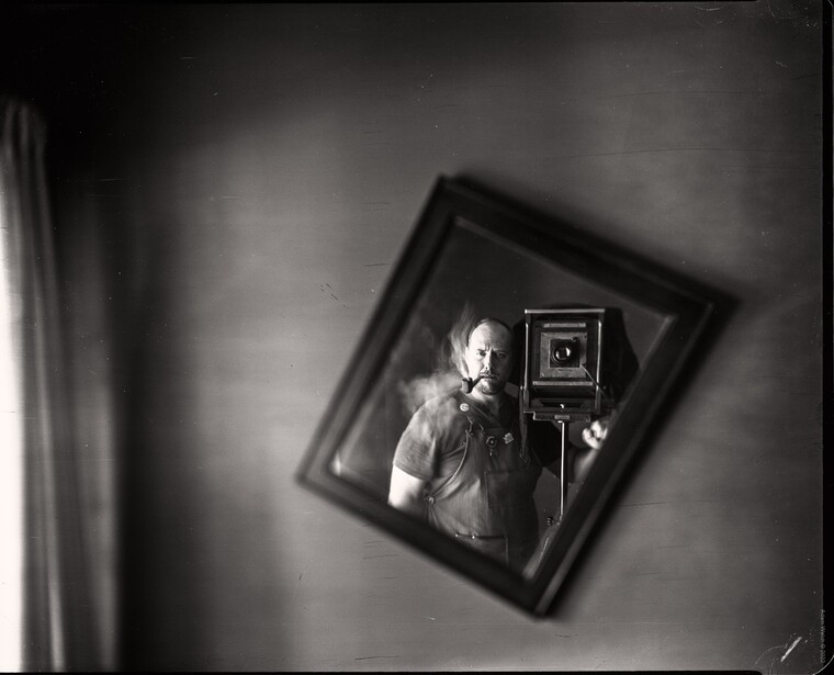 Black and white photograph showing a photographer with a large format camera on a tripod, viewed as a framed reflection or nested image.