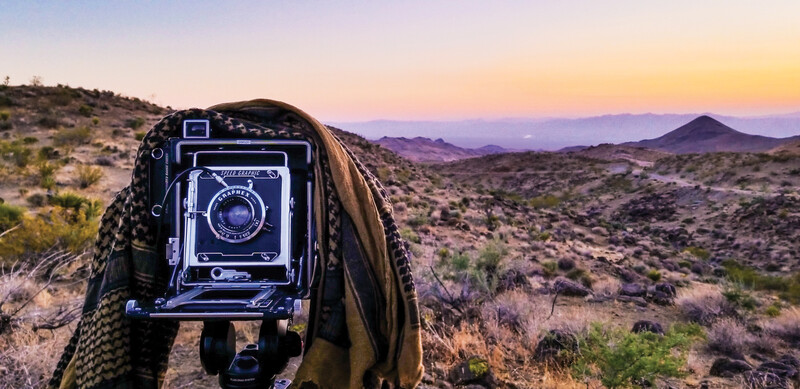 Large format camera held up to desert landscape at sunset with mountains in distance.
