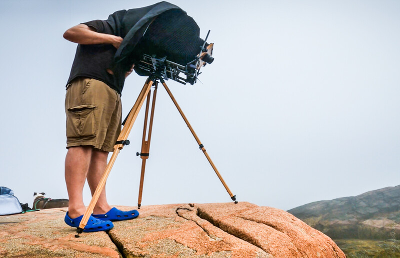 Cinematographer operating a film camera on a wooden tripod while standing on a rocky outcrop with a landscape vista behind.