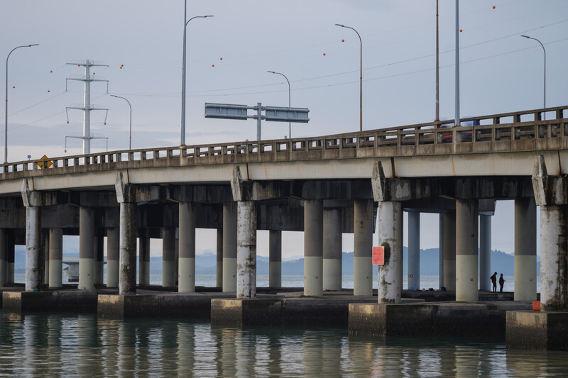 Concrete bridge with supporting pillars spanning calm water, with street lamps and directional sign overhead.