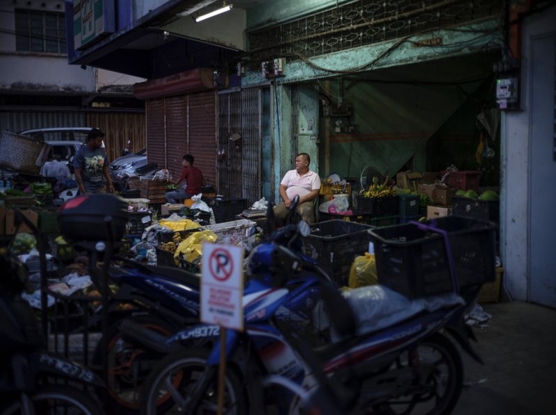 Man standing in cluttered street-level shop with motorcycles, merchandise, and tarps visible in warm, shadowed lighting.