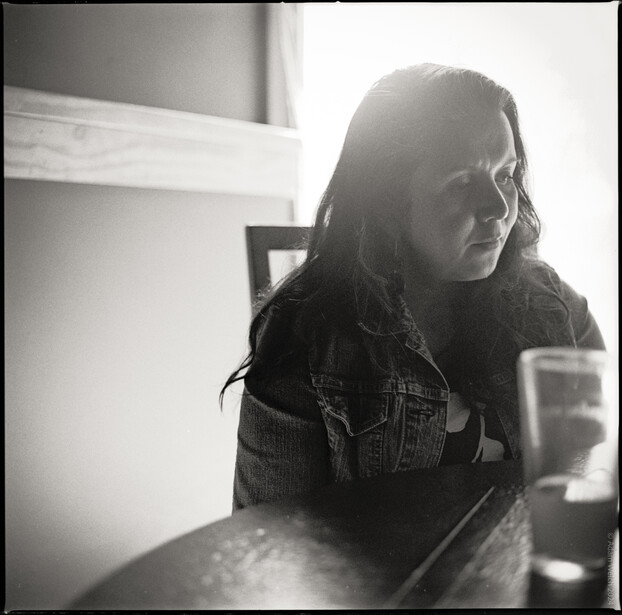 Woman with long dark hair wearing glasses and a denim jacket, photographed in profile against bright backlighting.