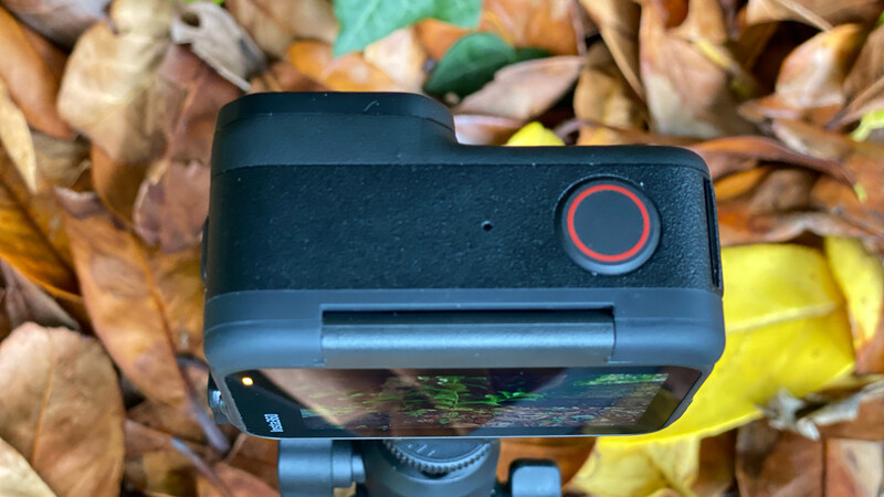 Action camera with red power button mounted on gimbal above autumn leaves.