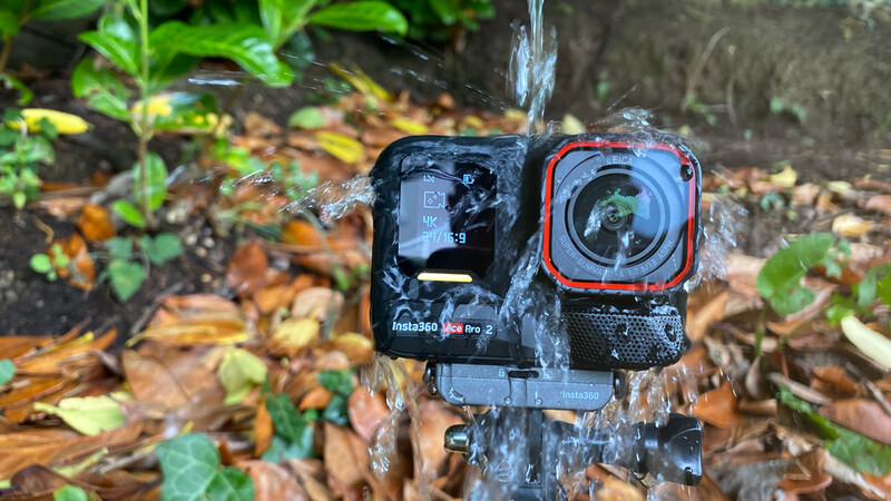 Two rugged action cameras mounted side-by-side on a log with water flowing over them in an outdoor forest setting.
