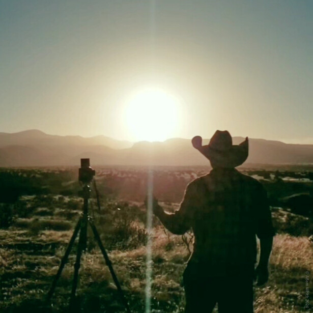 Photographer silhouetted against bright sunrise, operating camera on tripod in desert landscape.