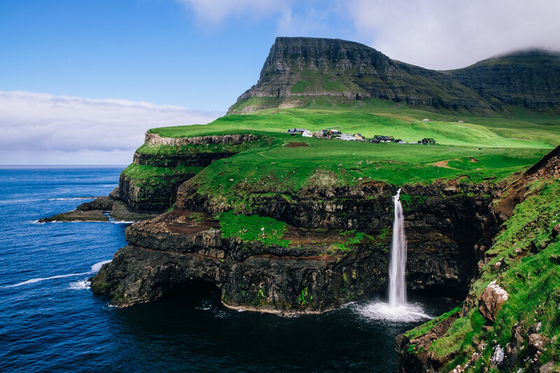 Waterfall cascading from a clifftop meadow into the ocean below, with green hillside and scattered buildings in the distance.