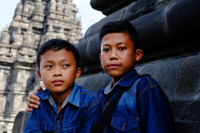 Two young boys in matching blue denim shirts posing against large stone blocks with an ornate temple structure visible in the background.
