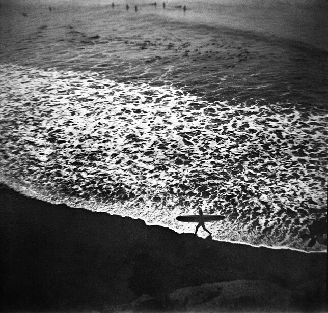 Black and white photograph of a wave breaking on a beach with figures visible in the water beyond.
