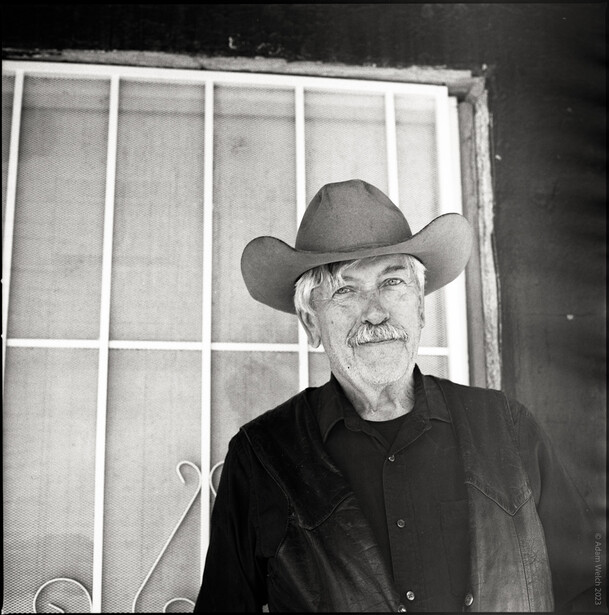 Older man wearing a cowboy hat and dark shirt, photographed indoors against a window with grid panes.