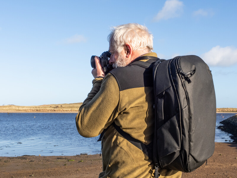 Photographer with white hair shooting by a coastal waterside with a large backpack and camera raised to eye level.