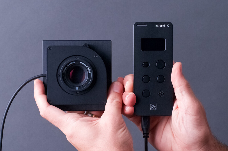 Hands holding a compact black camera module and its wireless remote control unit against a gray background.