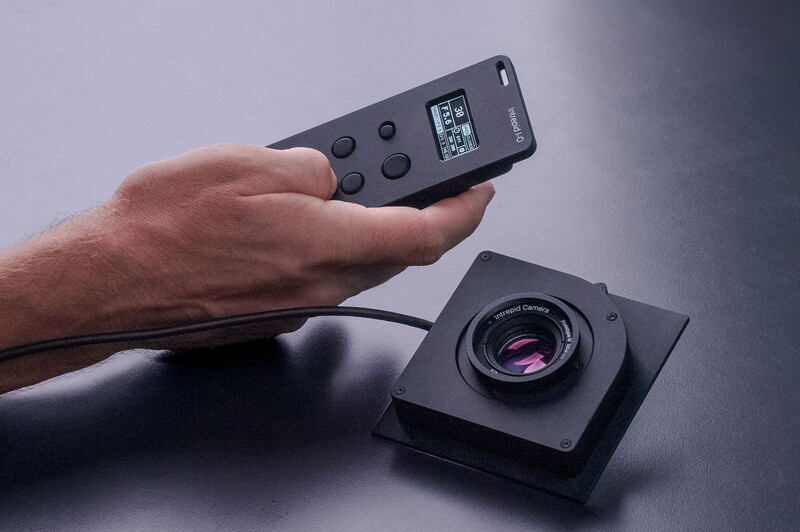 Hand holding wireless remote control above black camera lens mounted on gray surface.