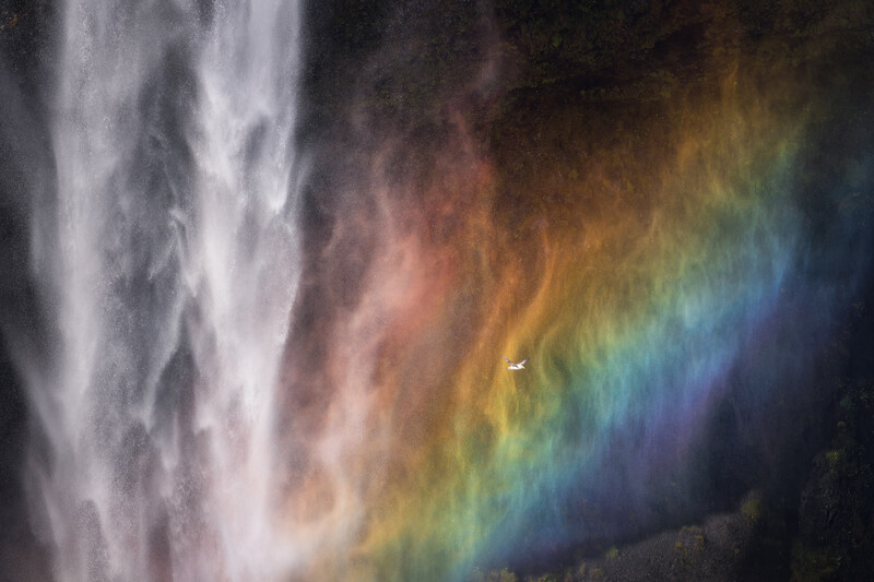 A vibrant rainbow arcing across a misty waterfall, with white water spray on the left side.