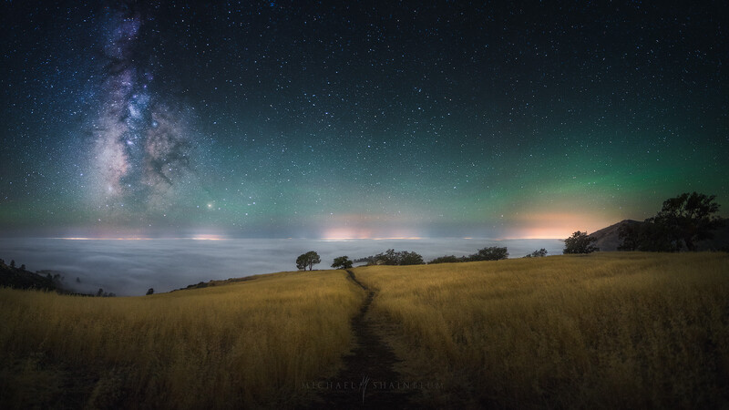 Milky Way arching over a misty valley landscape with a dirt path leading through golden grassland.