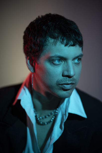 Man with short dark hair wearing a black jacket and light blue shirt with chain necklace, lit with contrasting red and blue gels.