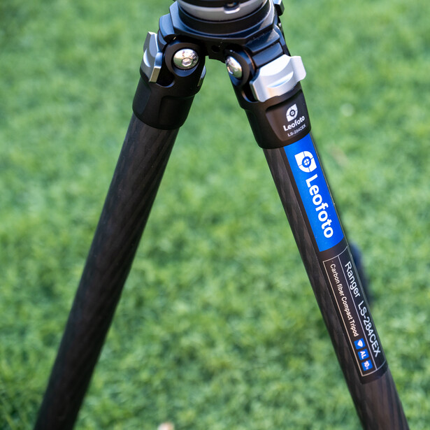 Close-up of a carbon fiber tripod leg with a blue brand label and adjustment mechanism against a blurred green background.