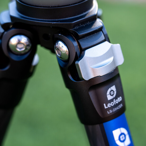 Close-up of a camera tripod head with ball joints and a blue leg clamp against a blurred green background.