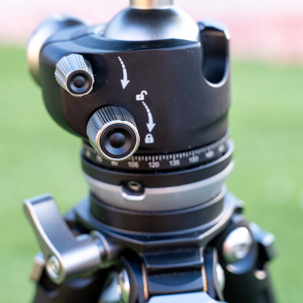 Close-up of a ball head tripod mount with dual adjustment knobs and graduated scale markings against a blurred outdoor background.