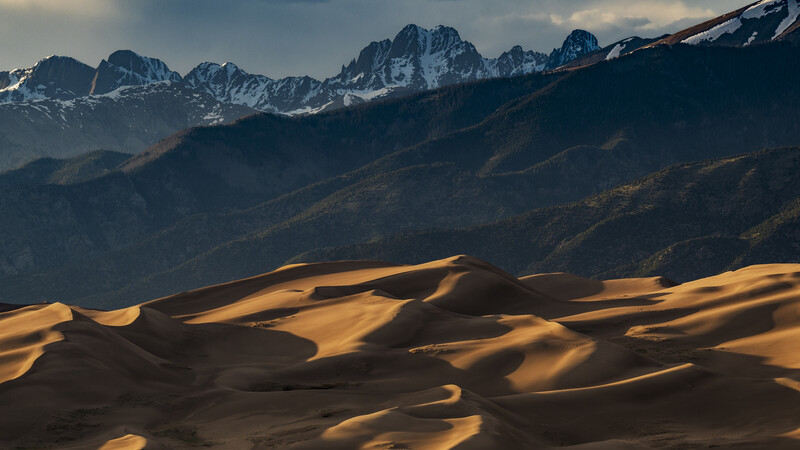 Golden sand dunes in foreground with snow-capped mountain range under blue sky in background.