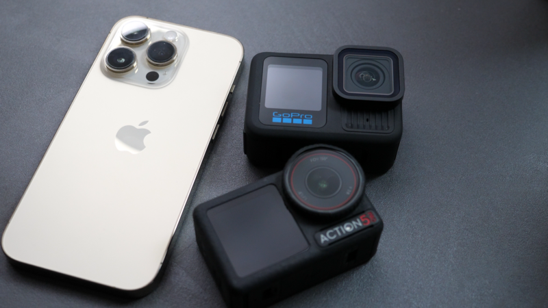 White iPhone 13 Pro positioned next to a black action camera and compact gimbal on gray surface.