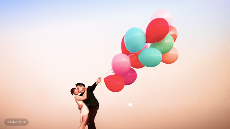 Couple in formal attire holding colorful balloons against a sunset sky.