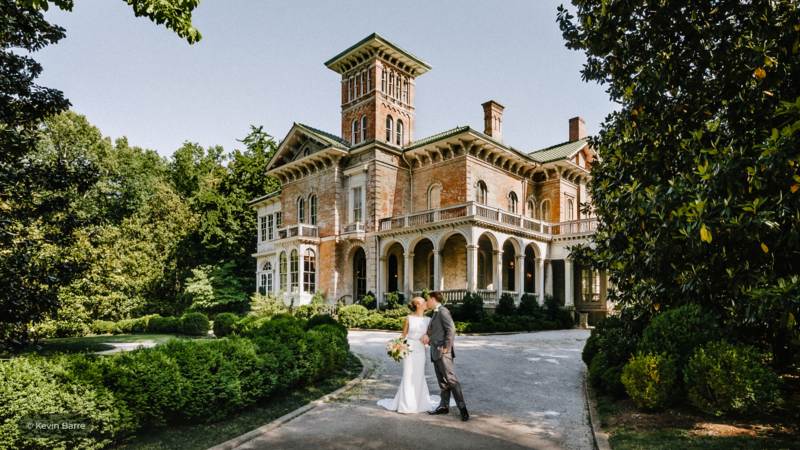 Bride and groom pose in front of a brick Victorian mansion with a tower and columned portico.