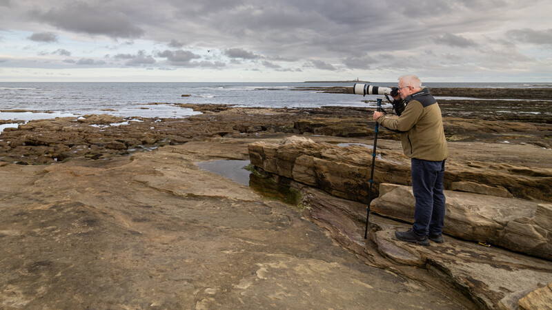 Photographer with telephoto lens mounted on tripod shooting seaward from rocky coastal outcrop.